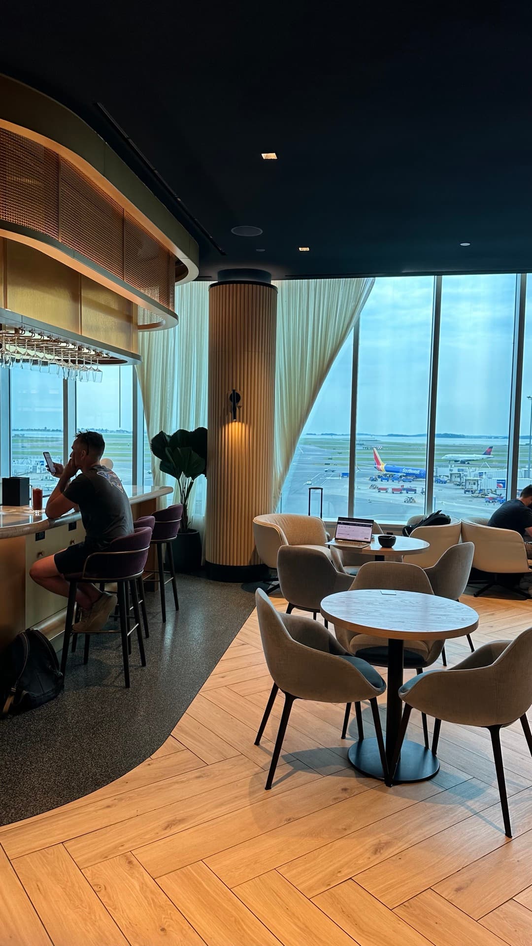 Interior of the Chase Sapphire Lounge in Terminal B at Boston Logan, with a bar, herringbone floor and floor-to-ceiling windows over the apron