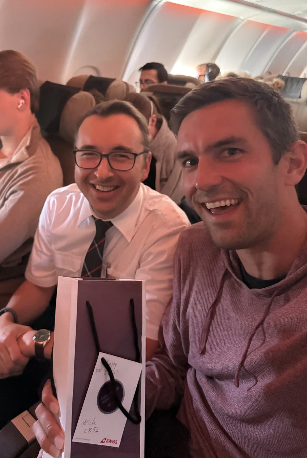 Jon Miksis and a SWISS flight attendant smiling in the cabin, holding a gift bag with a handwritten note